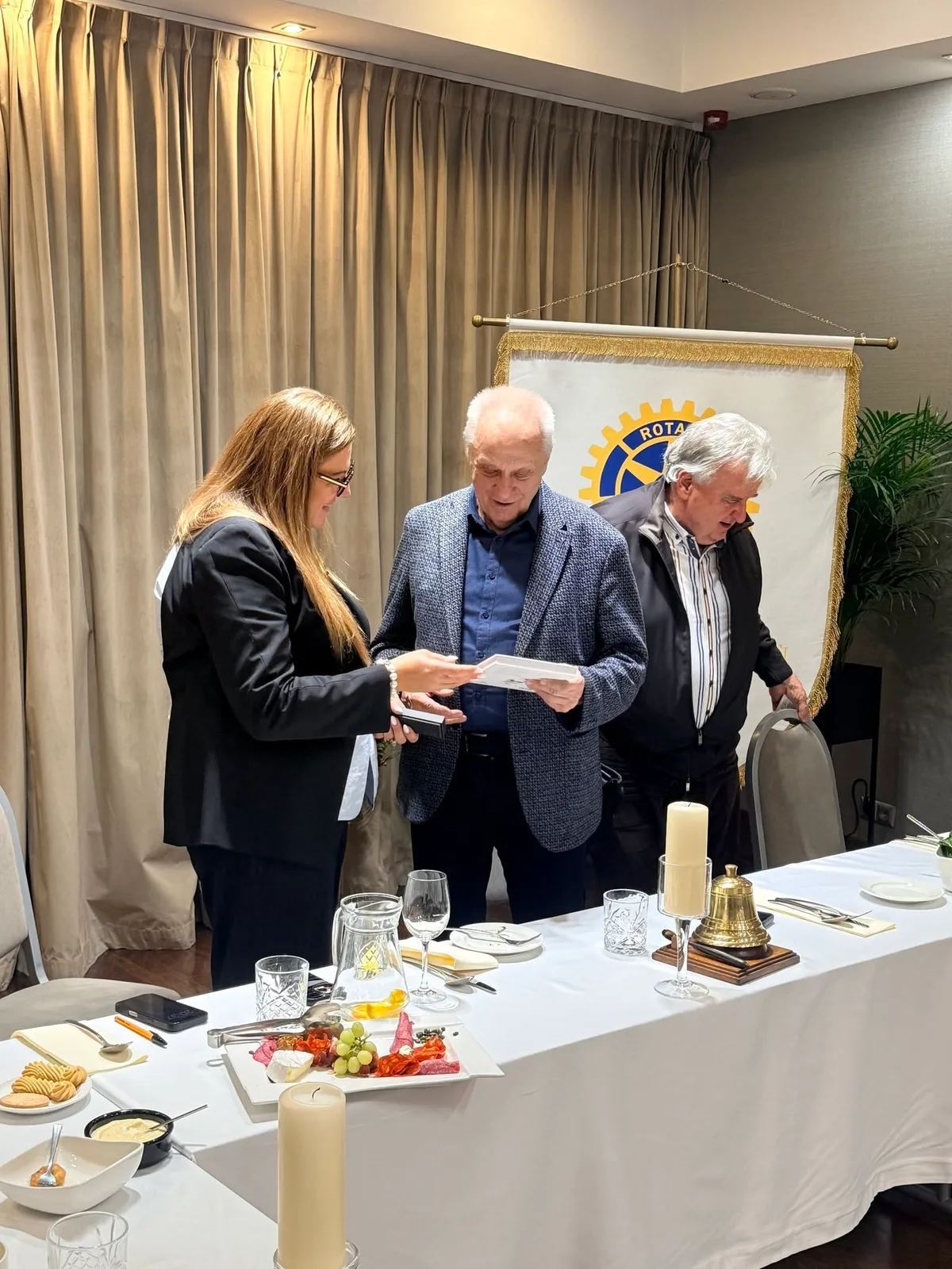 Kobieta wręcza gościowi spotkania upominek i dokument, obok stoi drugi mężczyzna, w tle proporczyk Rotary w sali hotelowej.