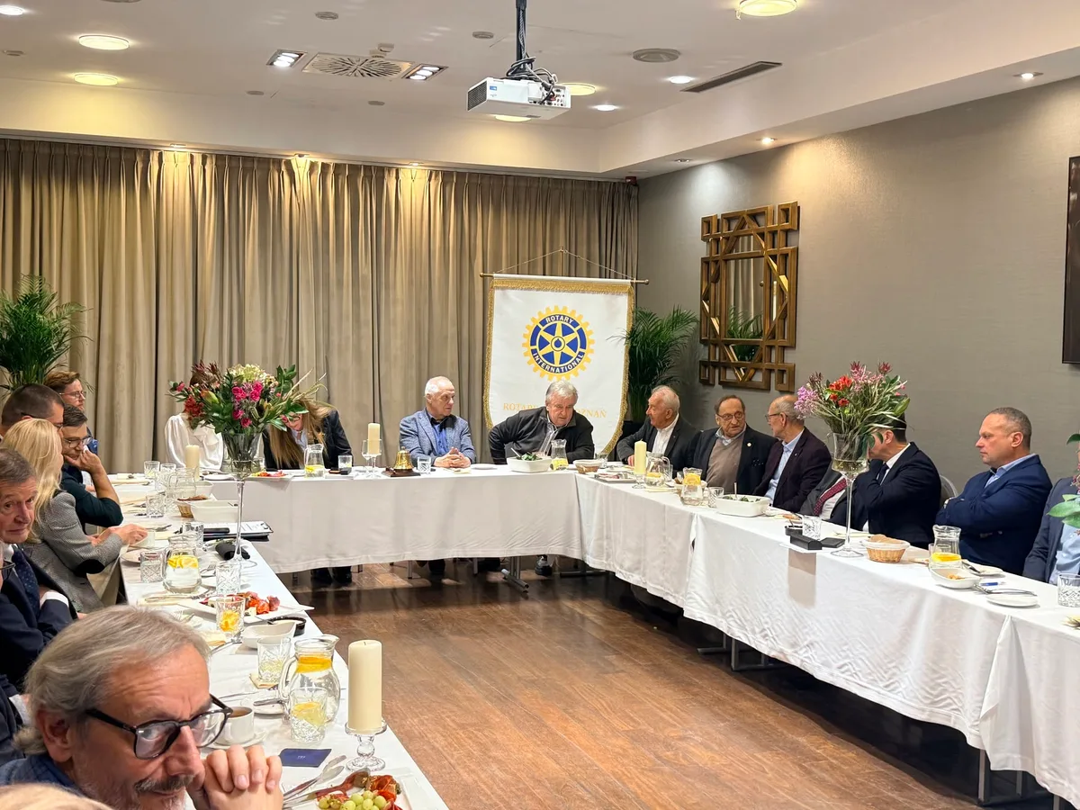 Członkowie klubu siedzą przy stołach w kształcie litery U w sali hotelowej, w głębi chorągiewka Rotary International.