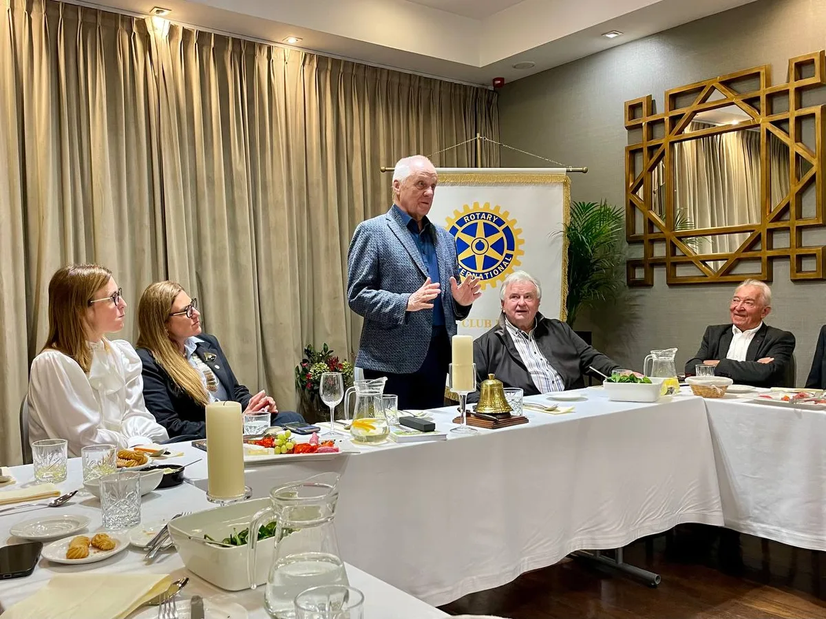 Gość spotkania stoi i przemawia przy stole, za nim baner Rotary International, obok siedzą członkowie klubu.